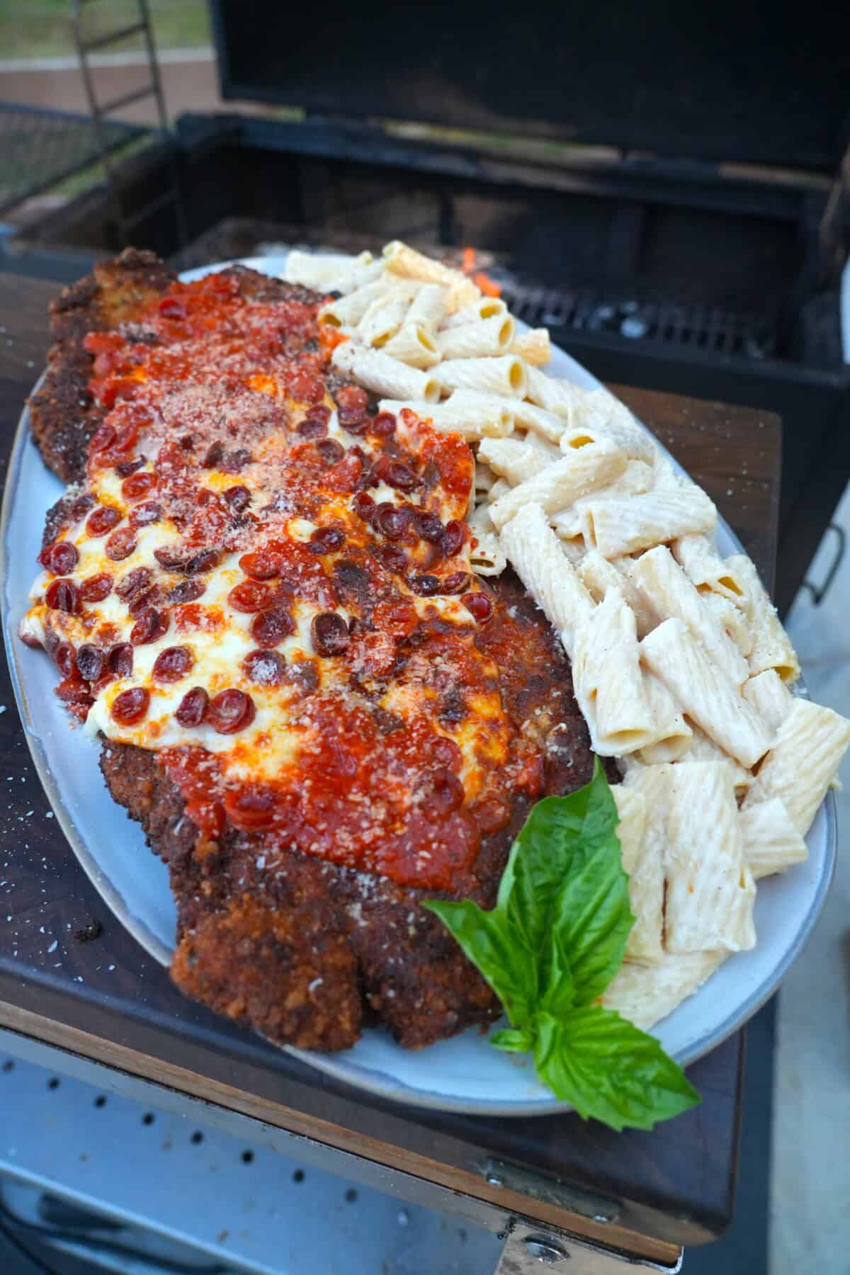 A plate of chicken pizza Parmesan with pepperoni, marinara sauce and cheese on top of the breaded chicken, with basil garnish and rigatoni Alfredo on the side