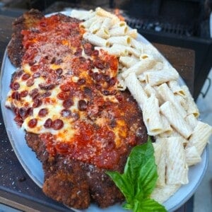 A plate of chicken pizza Parmesan with pepperoni, marinara sauce and cheese on top of the breaded chicken, with basil garnish and rigatoni Alfredo on the side
