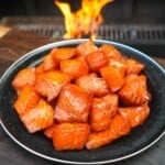 A finished plate of maple bourbon salmon burnt ends, with a grill fire in the background