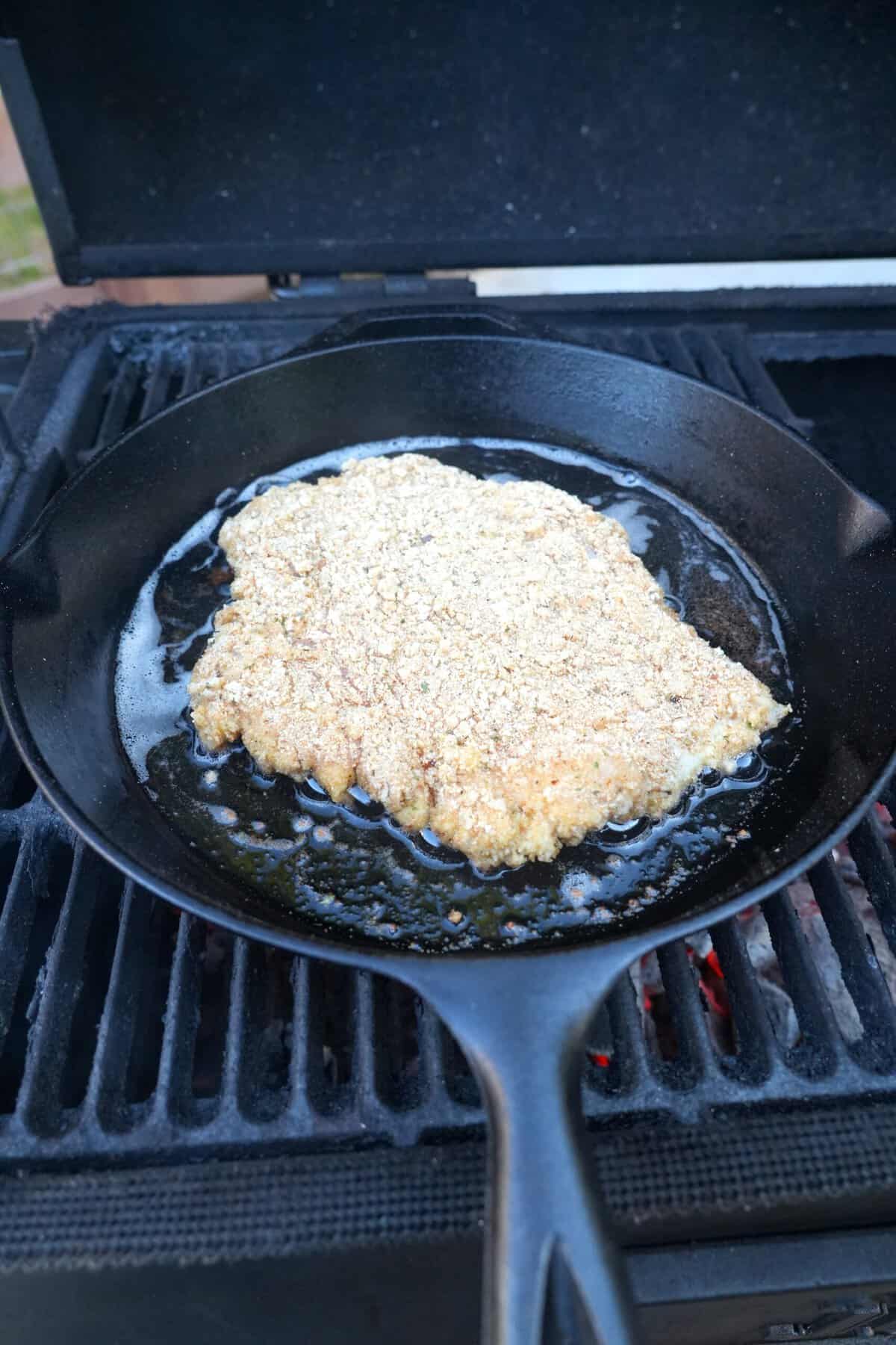 Breaded chicken cutlets frying in olive oil in a cast iron skillet on the grill