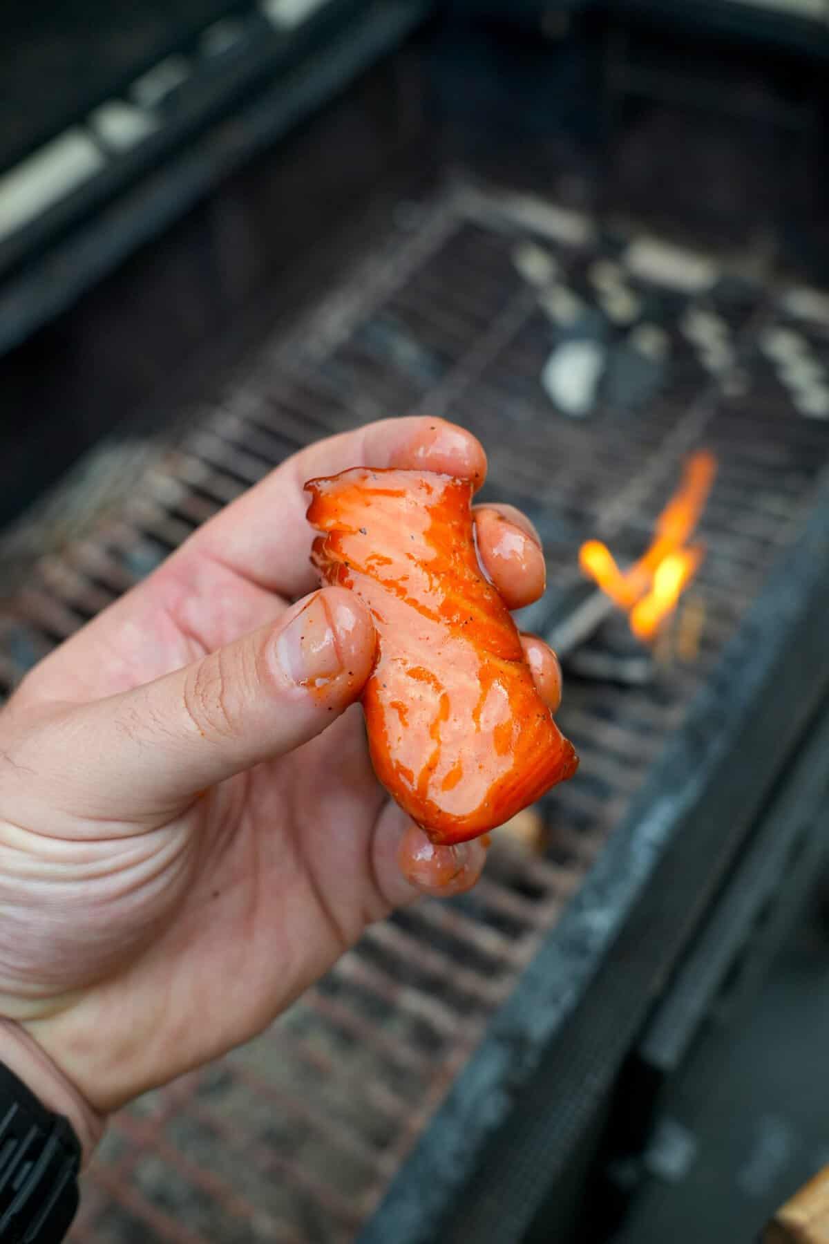Derek Wolf holding up a maple smoked salmon burnt end, with the smoker and fire in the background
