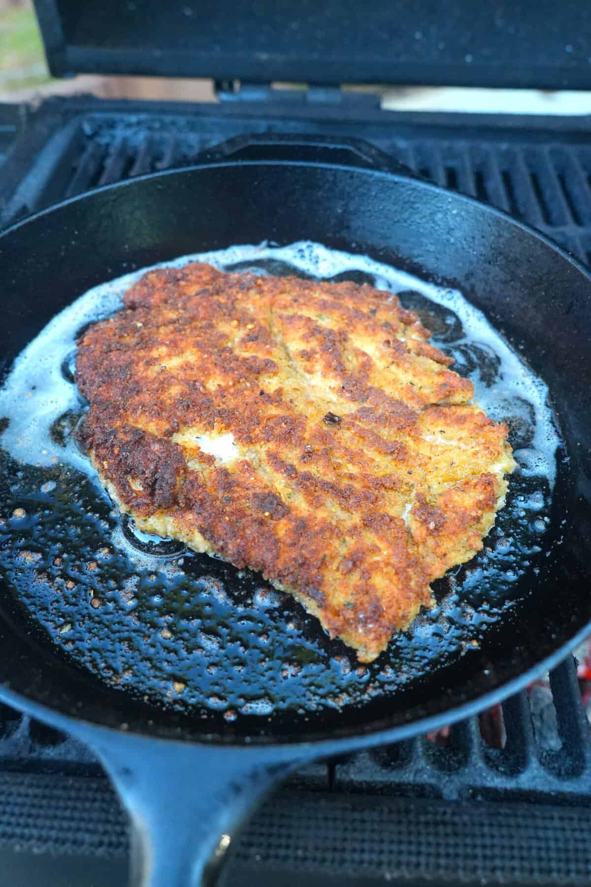 A finished chicken Parmesan cutlet with a golden brown crust, sitting in olive oil in a cast iron skillet