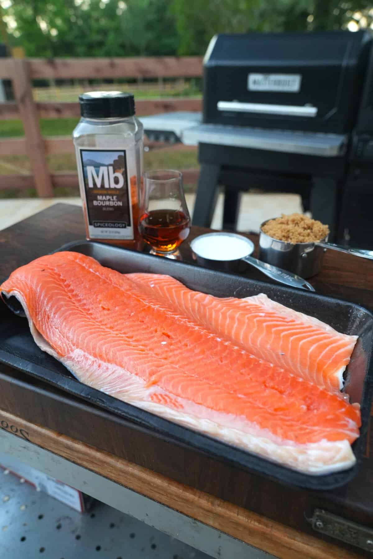A raw side of salmon on a cutting board, with Maple Bourbon Seasoning and other curing and glazing ingredients in the background