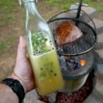 Derek Wolf holding a glass bottle full of beer salmuera, with a grilled steak on a charcoal grill in the background