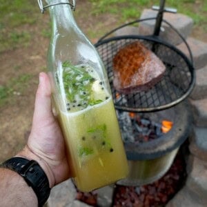 Derek Wolf holding a glass bottle full of beer salmuera, with a grilled steak on a charcoal grill in the background