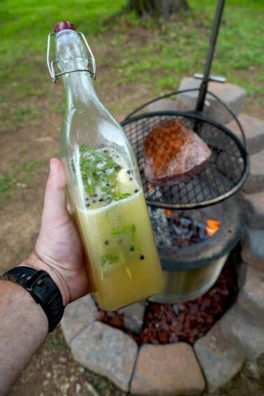Derek Wolf holding a glass bottle full of beer salmuera, with a grilled steak on a charcoal grill in the background