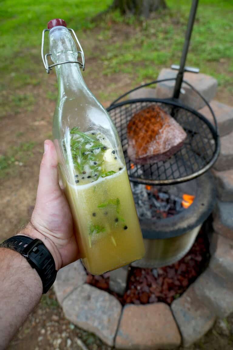 Derek Wolf holding a glass bottle full of beer salmuera, with a grilled steak on a charcoal grill in the background