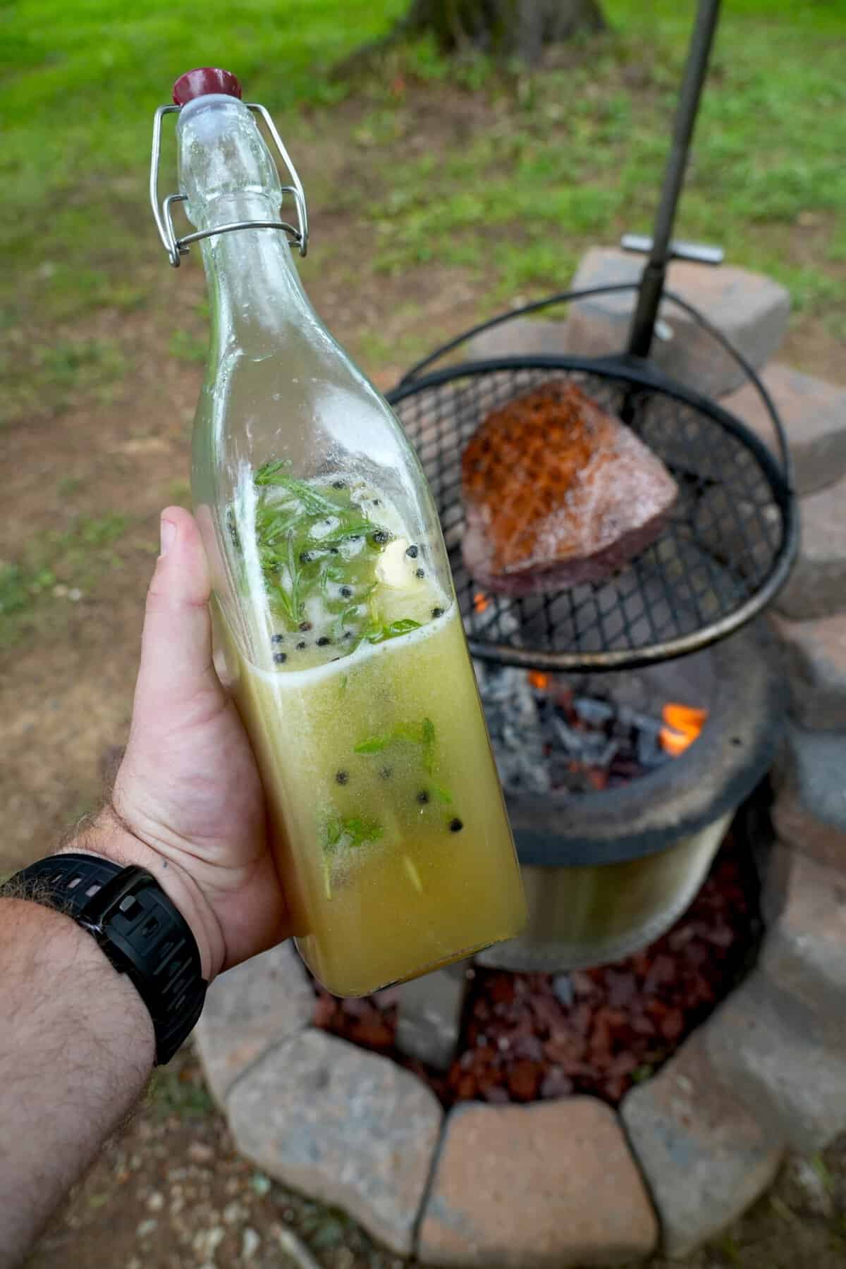 Derek Wolf holding a glass bottle full of beer salmuera, with a grilled steak on a charcoal grill in the background