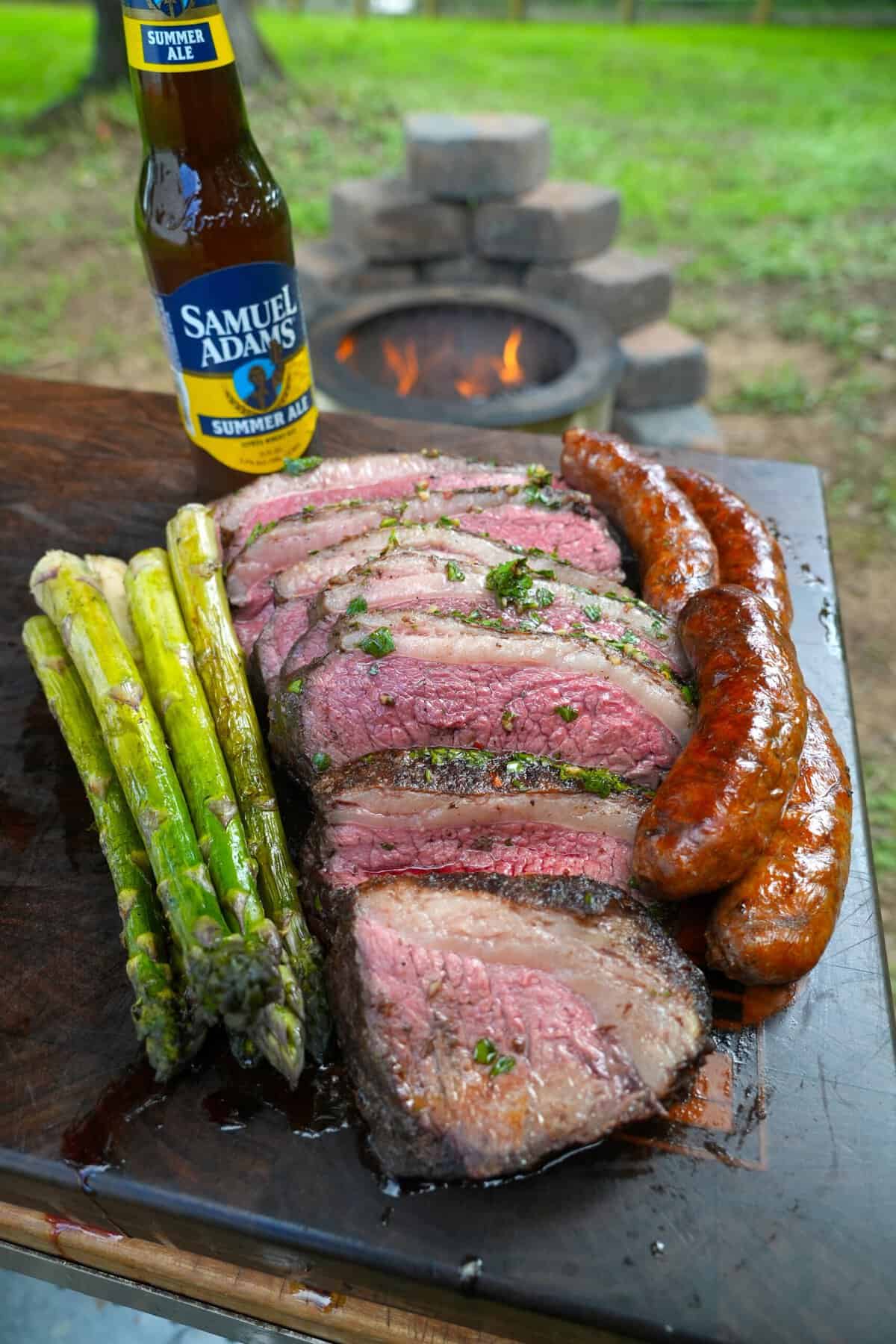 Another shot of grilled, sliced steak, cooked sausages and grilled asparagus on a cutting board with a bottle of Samuel Adams in the background