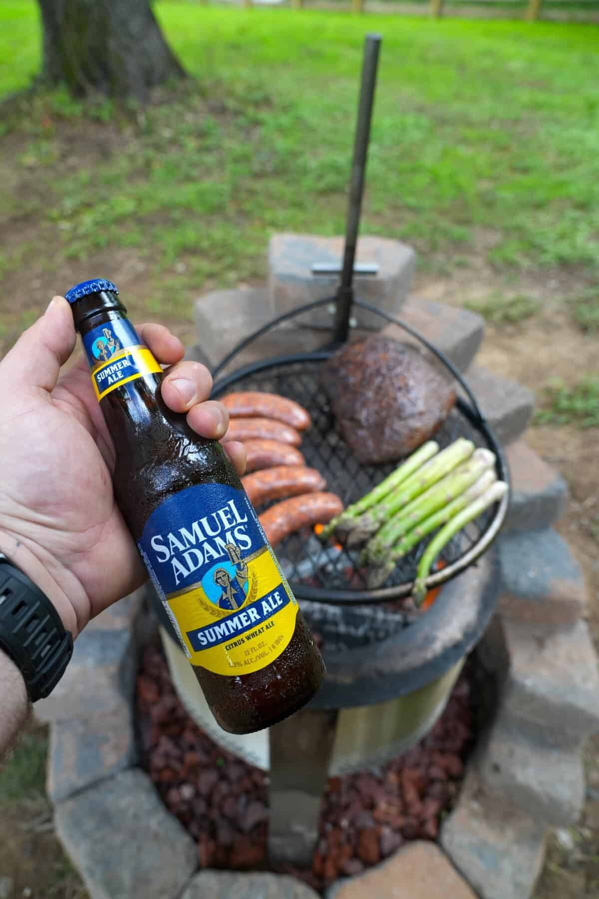 Derek Wolf holding up a bottle of Samuel Adams Summer Ale, in front of grilled steak and asparagus on a charcoal grill