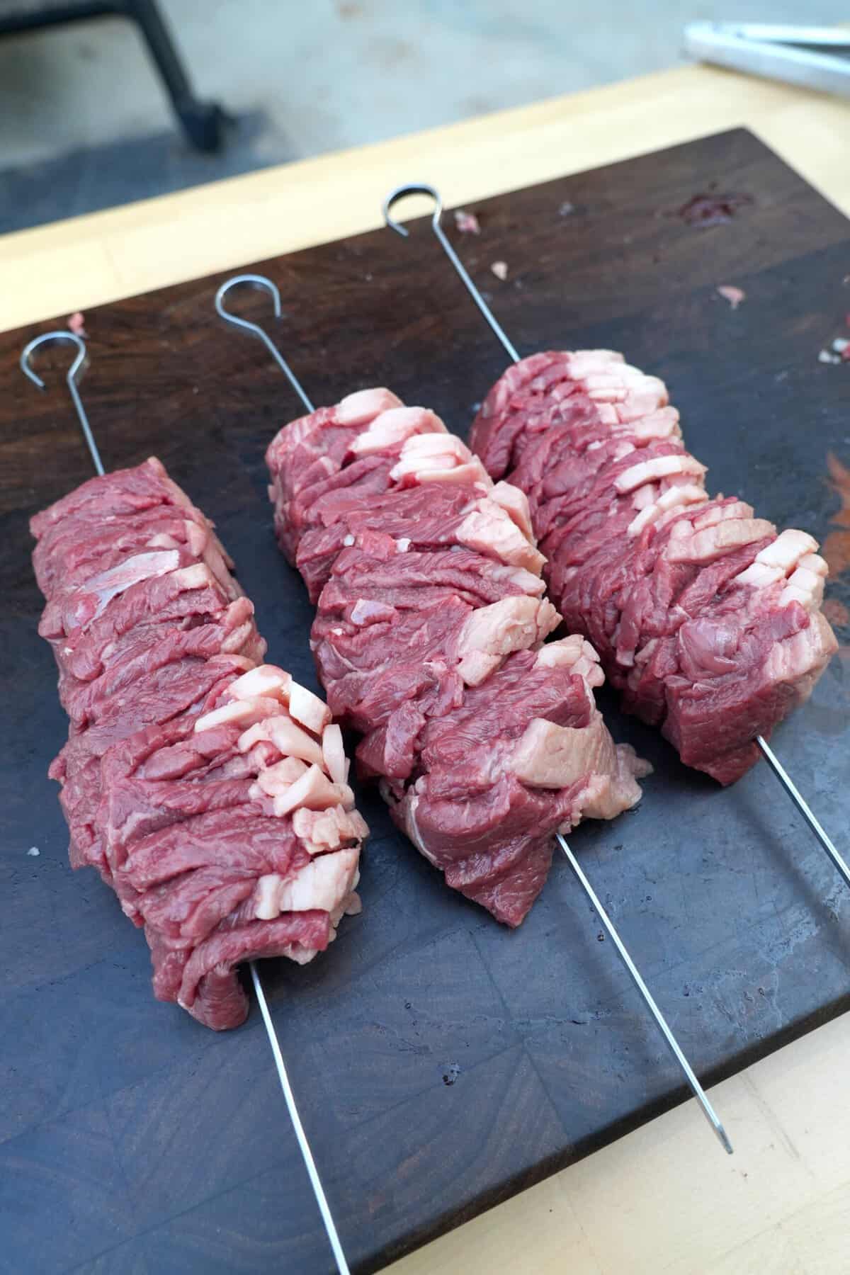 Raw picanha steak skewers ready for grilling on a black cutting board.