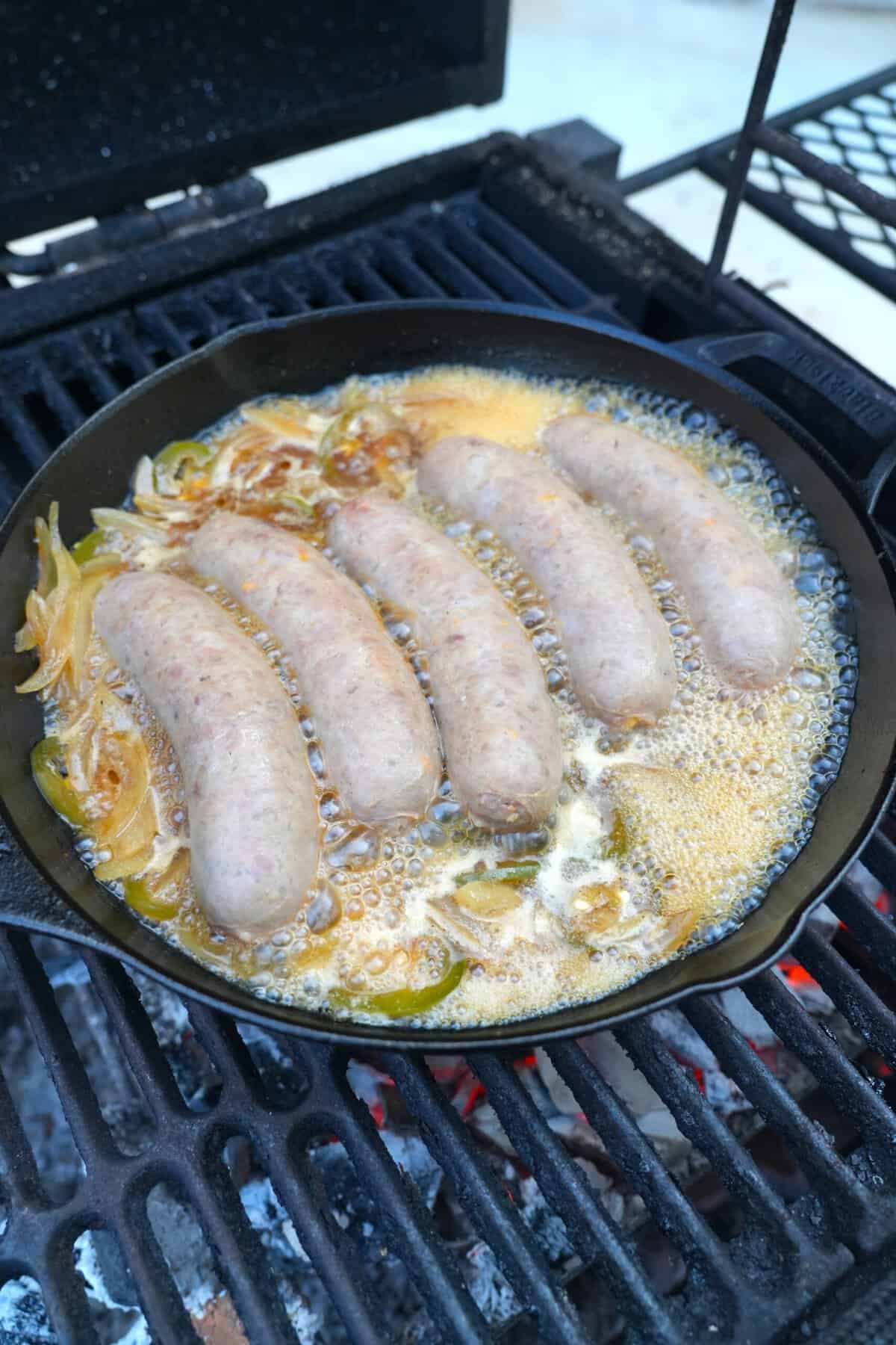 Five beer brats simmering in Samuel Adams Octoberfest in a cast iron skillet on grill grates