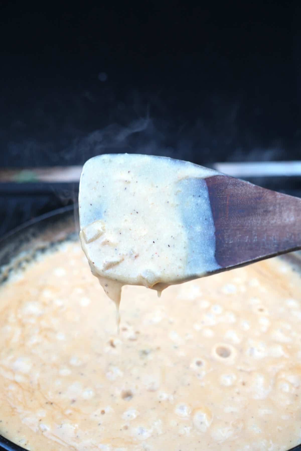 A wooden spoon coated in beer cheese to show texture, held above a cast iron skillet full of bubbling beer cheese