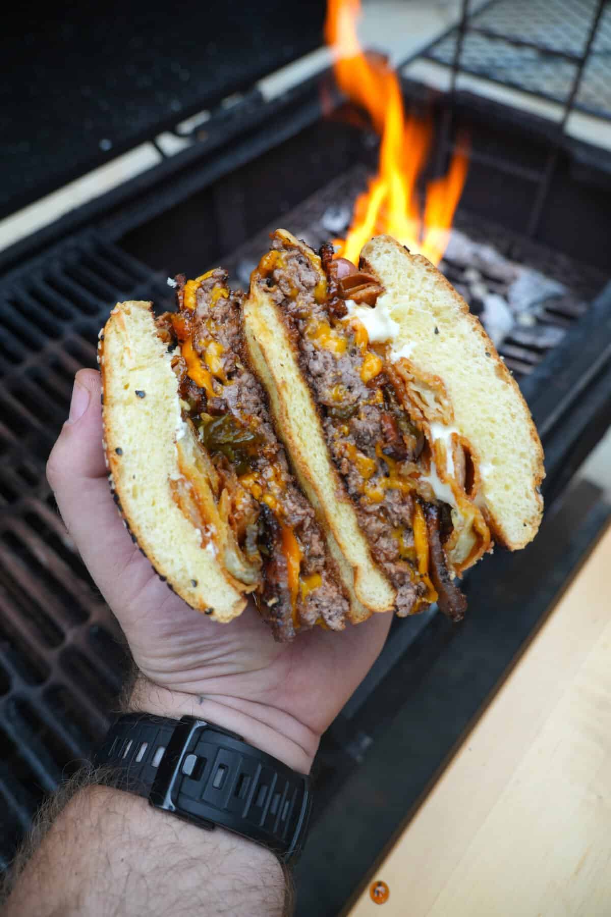 A close up of the Western BBQ Burger, cut in half, held in Derek Wolf's hand, with fire from the grill flaming in the background.