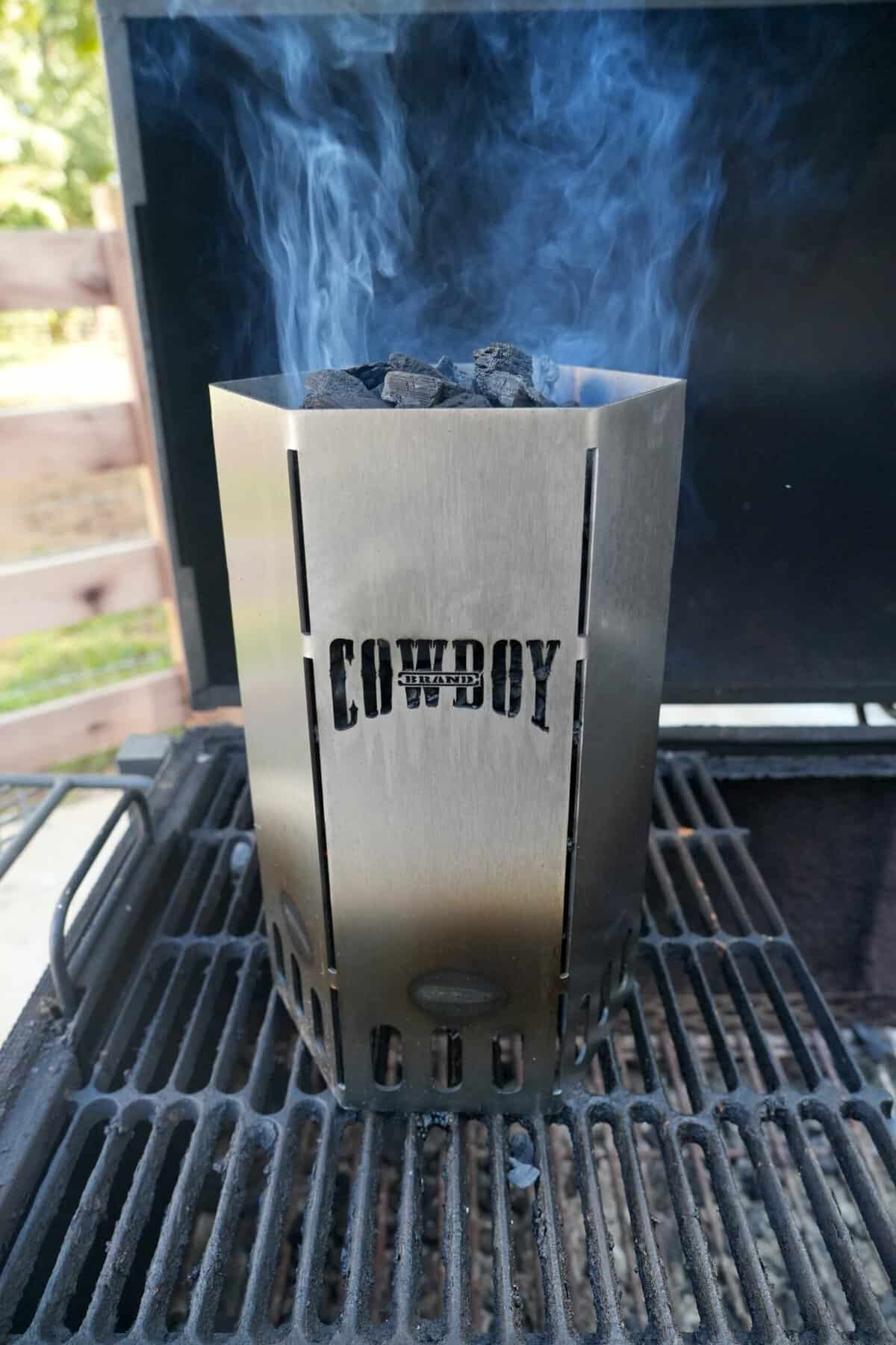 A branded Cowboy Charcoal chimney starter, smoking on top of grill grates