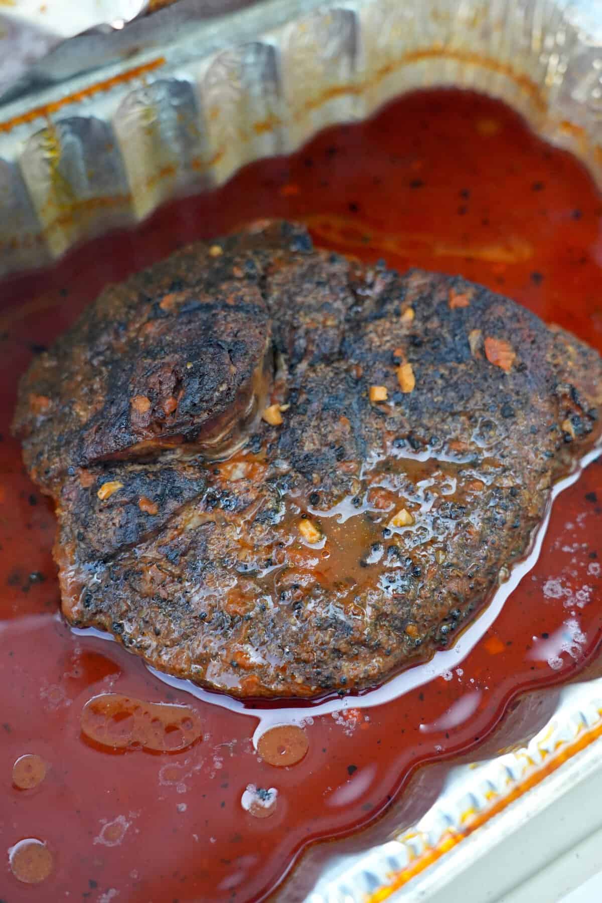 A close up of a cooked grilled chuck roast, sitting in braising liquid in the foil pan
