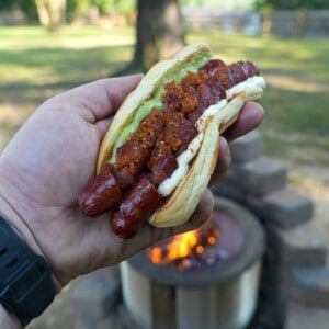 Derek Wolf holding a finished spiral hot dog in a bun with guacamole, mayonnaise, mustard and pico de gallo, with a fire pit and the woods in the background