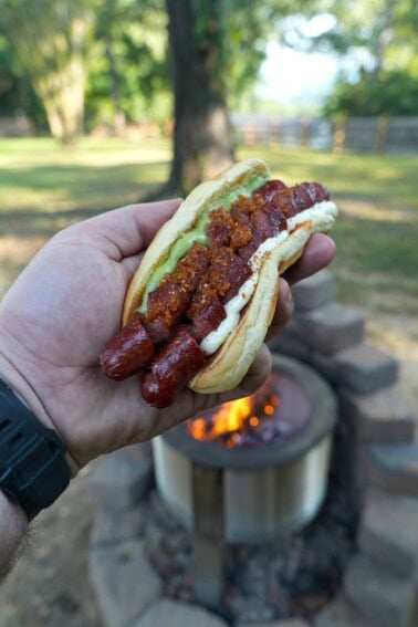 Derek Wolf holding a finished spiral hot dog in a bun with guacamole, mayonnaise, mustard and pico de gallo, with a fire pit and the woods in the background