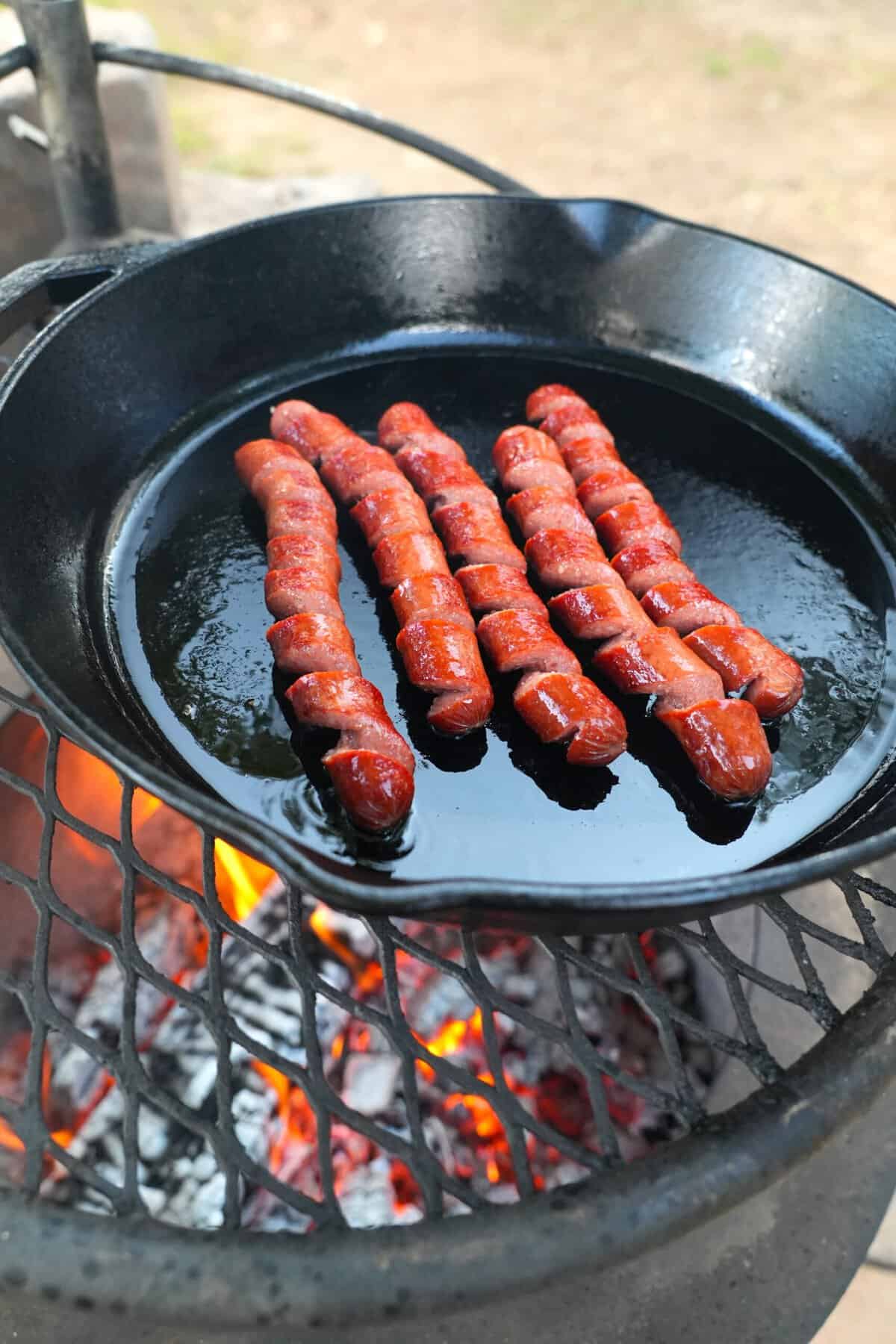 Five spiral cut hot dogs cooking in a skillet over a fire pit
