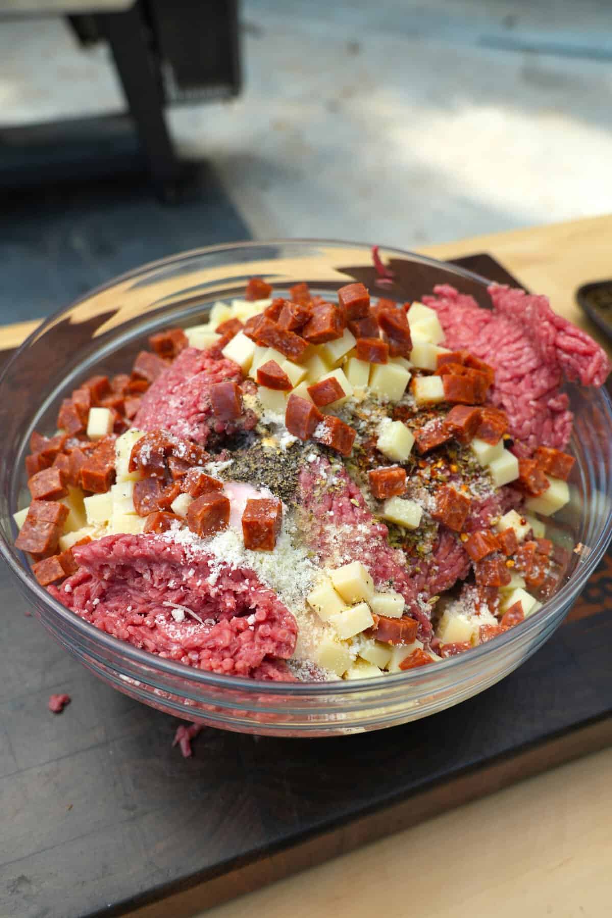 Ingredients for pizza beef sticks, including ground beef, pepperoni, cheese and Italian seasonings, in a glass bowl on a cutting board