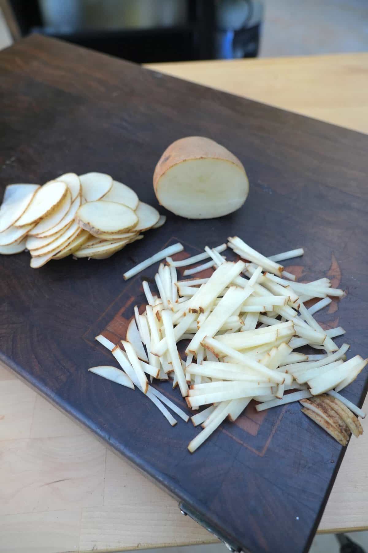 A pile of raw shoestring fries cut on a cutting board, with slices of uncut potato to the left and half of a potato (not sliced) to the right