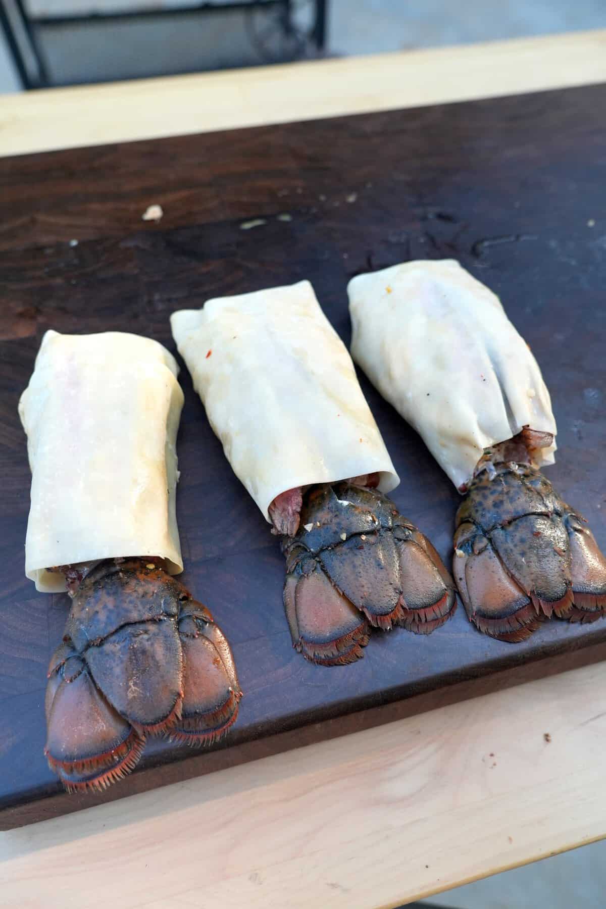 Three uncooked lobster tails wrapped in egg roll wrappers on a cutting board