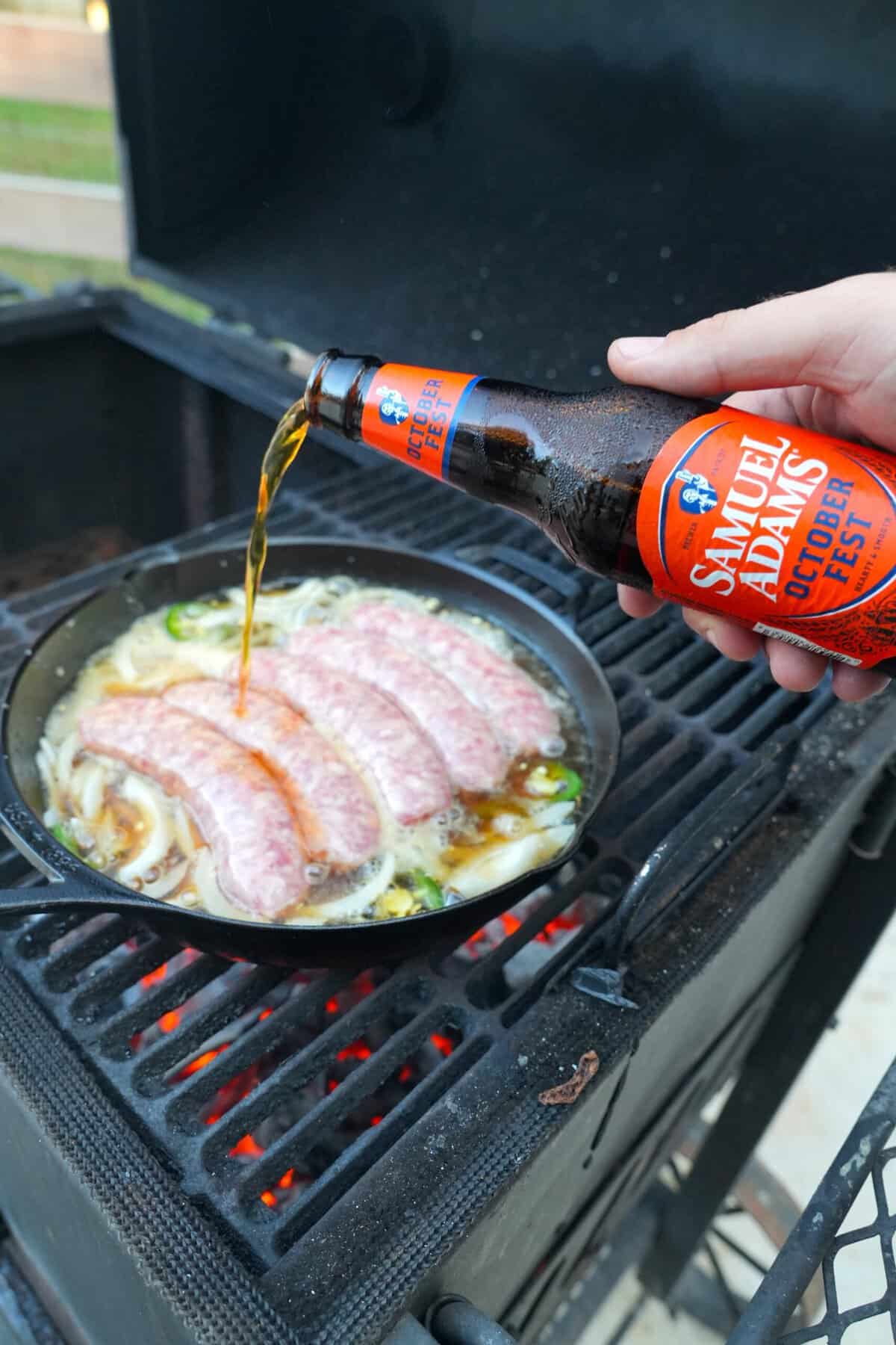 Derek Wolf pouring a bottle of Samuel Adams Octoberfest in a cast iron skillet, which has five beer brats and onions and jalapeños inside