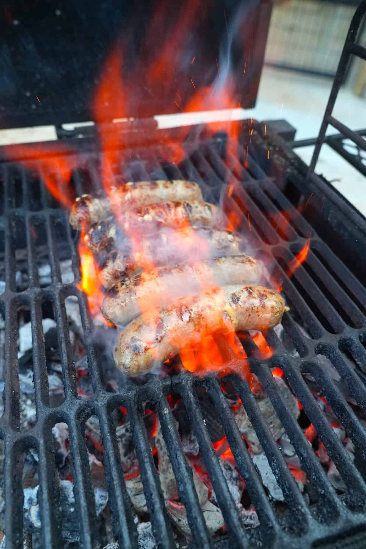 Five beer brats searing on a hot grill, with red flames coming up around them