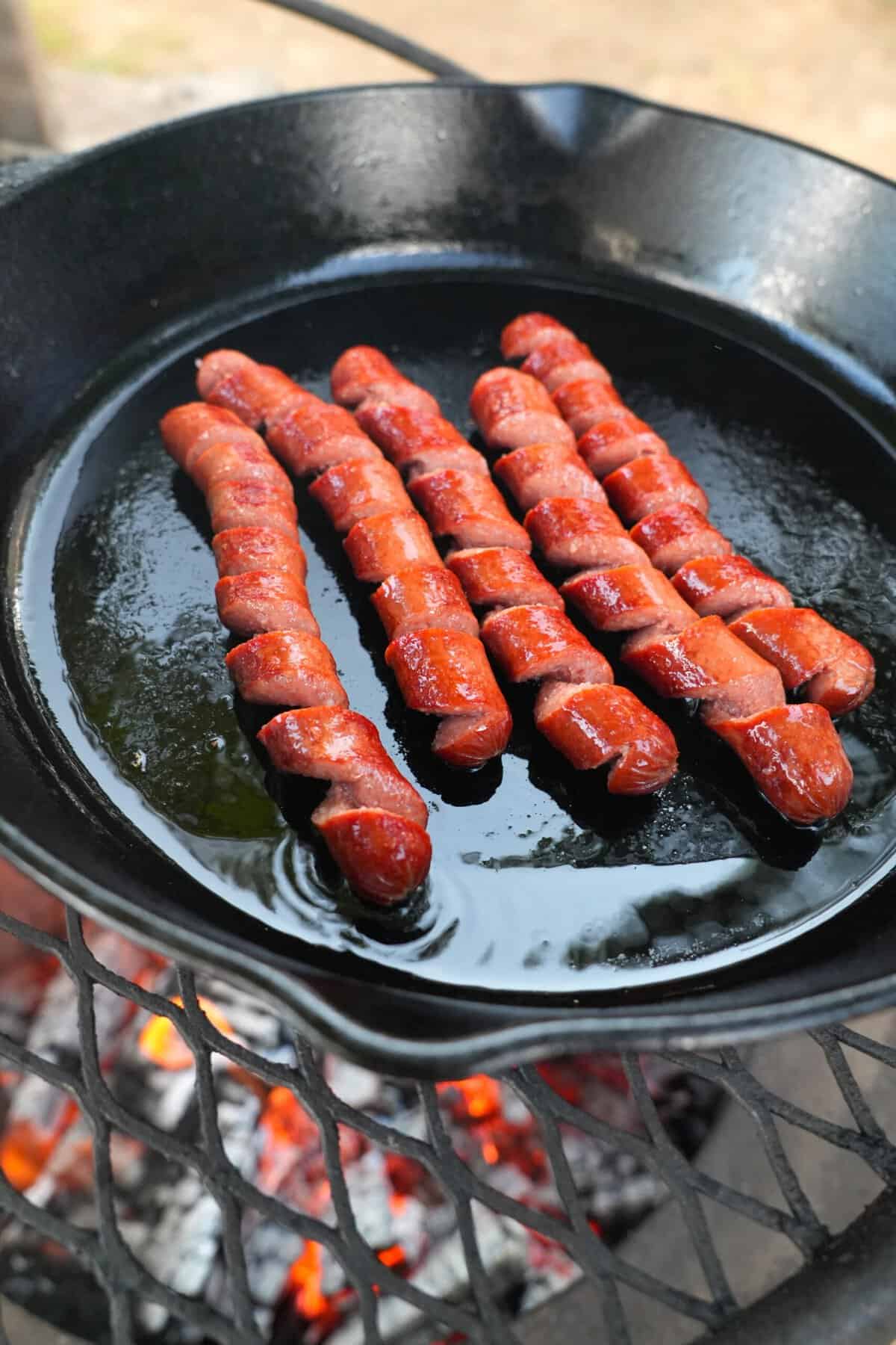A close up of five spiral cut hot dogs cooking on a greased cast iron skillet over the fire pit