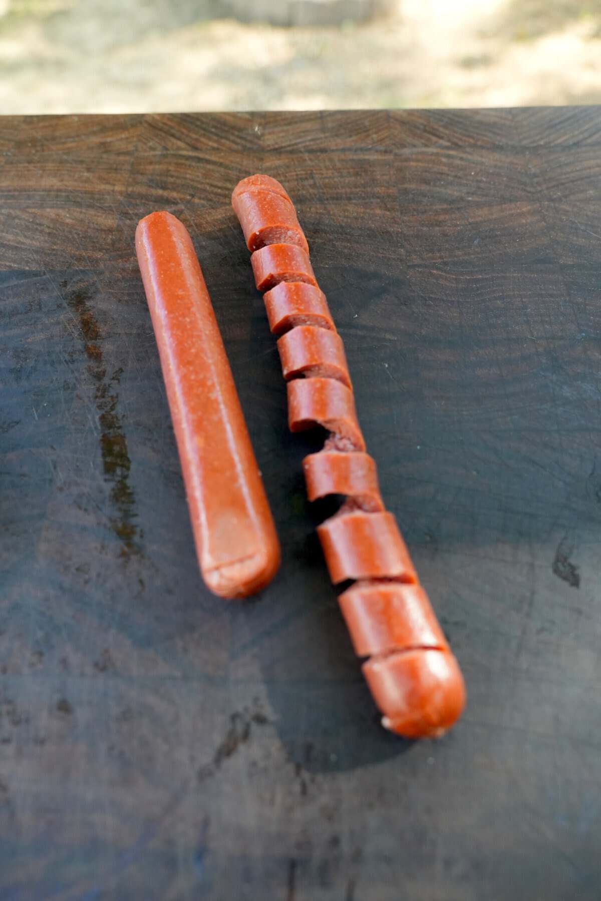An uncut hot dog lying next to a spiral cut, stretched hot dog on a cutting board