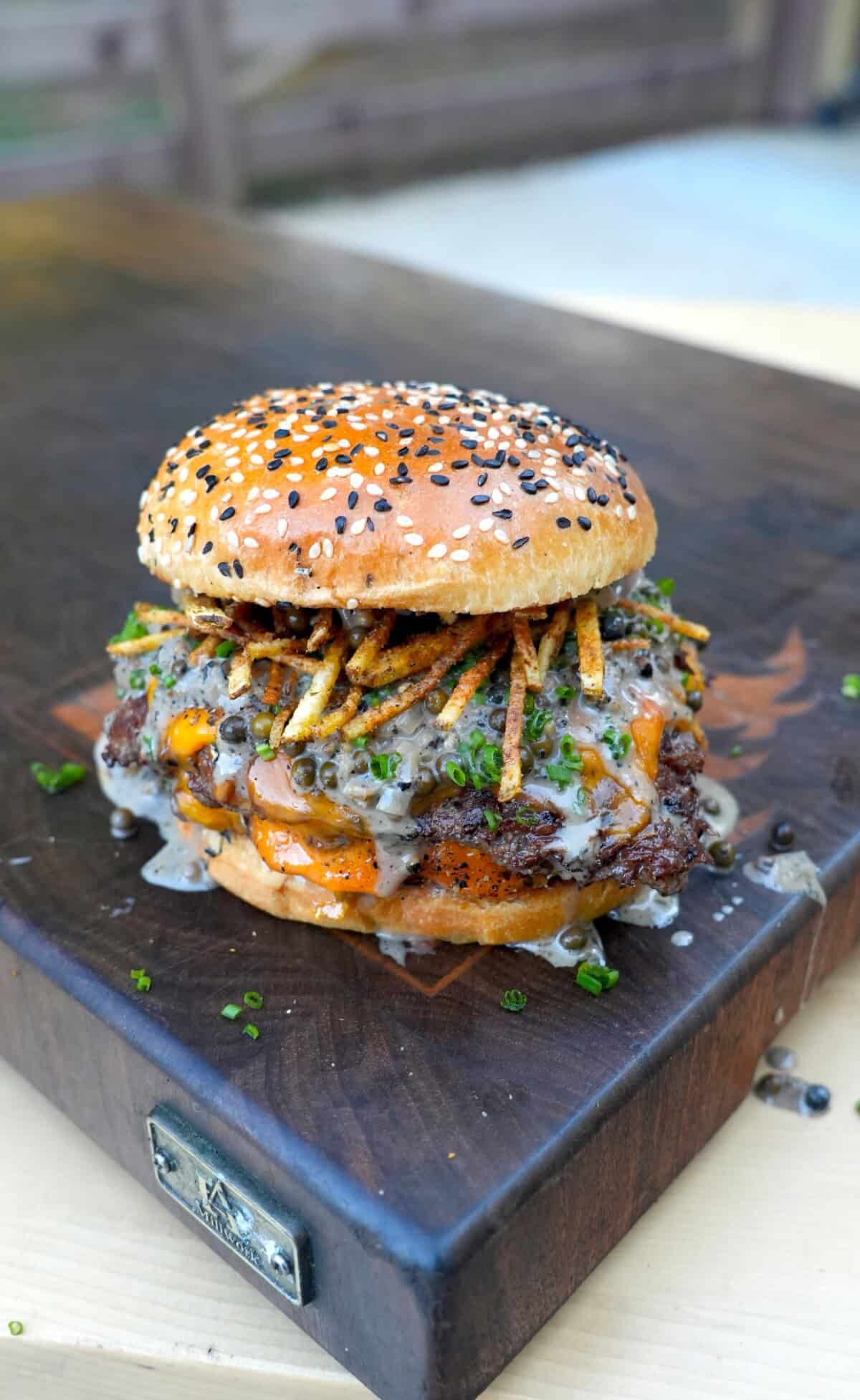 A fully assembled steak au poivre burger on a cutting board, with shoestring fries and whisky peppercorn sauce on the patty sitting inside a toasted everything seasoned bun
