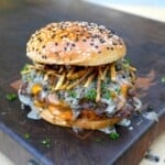 A fully assembled steak au poivre burger on a cutting board, with shoestring fries and whisky peppercorn sauce on the patty sitting inside a toasted everything seasoned bun