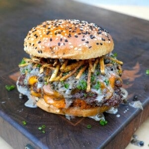 A fully assembled steak au poivre burger on a cutting board, with shoestring fries and whisky peppercorn sauce on the patty sitting inside a toasted everything seasoned bun