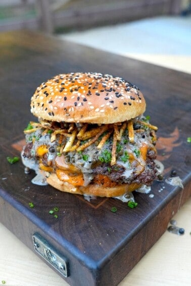 A fully assembled steak au poivre burger on a cutting board, with shoestring fries and whisky peppercorn sauce on the patty sitting inside a toasted everything seasoned bun