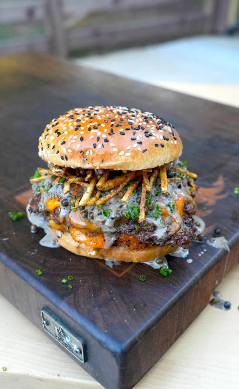 A fully assembled steak au poivre burger on a cutting board, with shoestring fries and whisky peppercorn sauce on the patty sitting inside a toasted everything seasoned bun