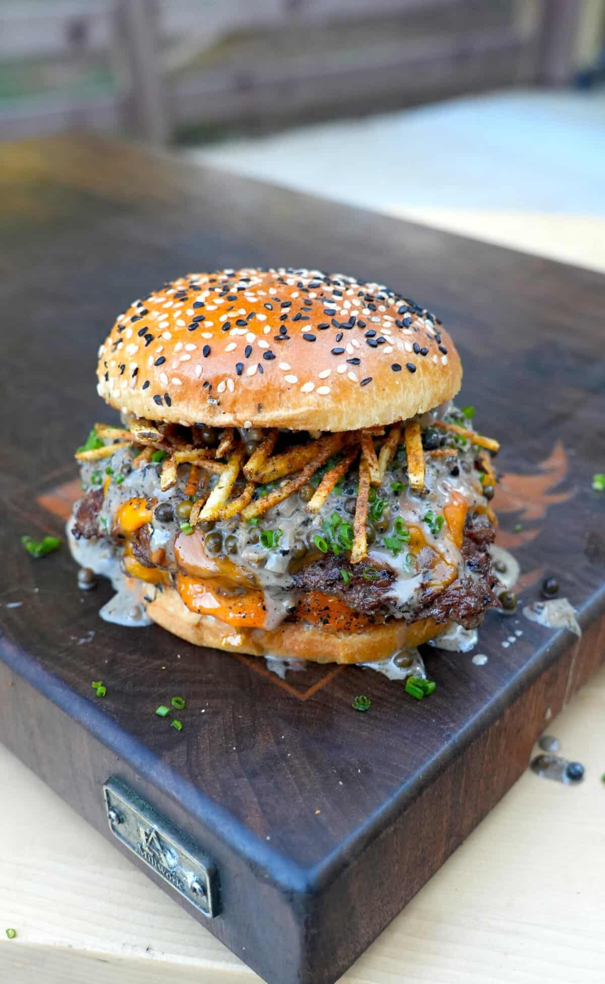 A fully assembled steak au poivre burger on a cutting board, with shoestring fries and whisky peppercorn sauce on the patty sitting inside a toasted everything seasoned bun