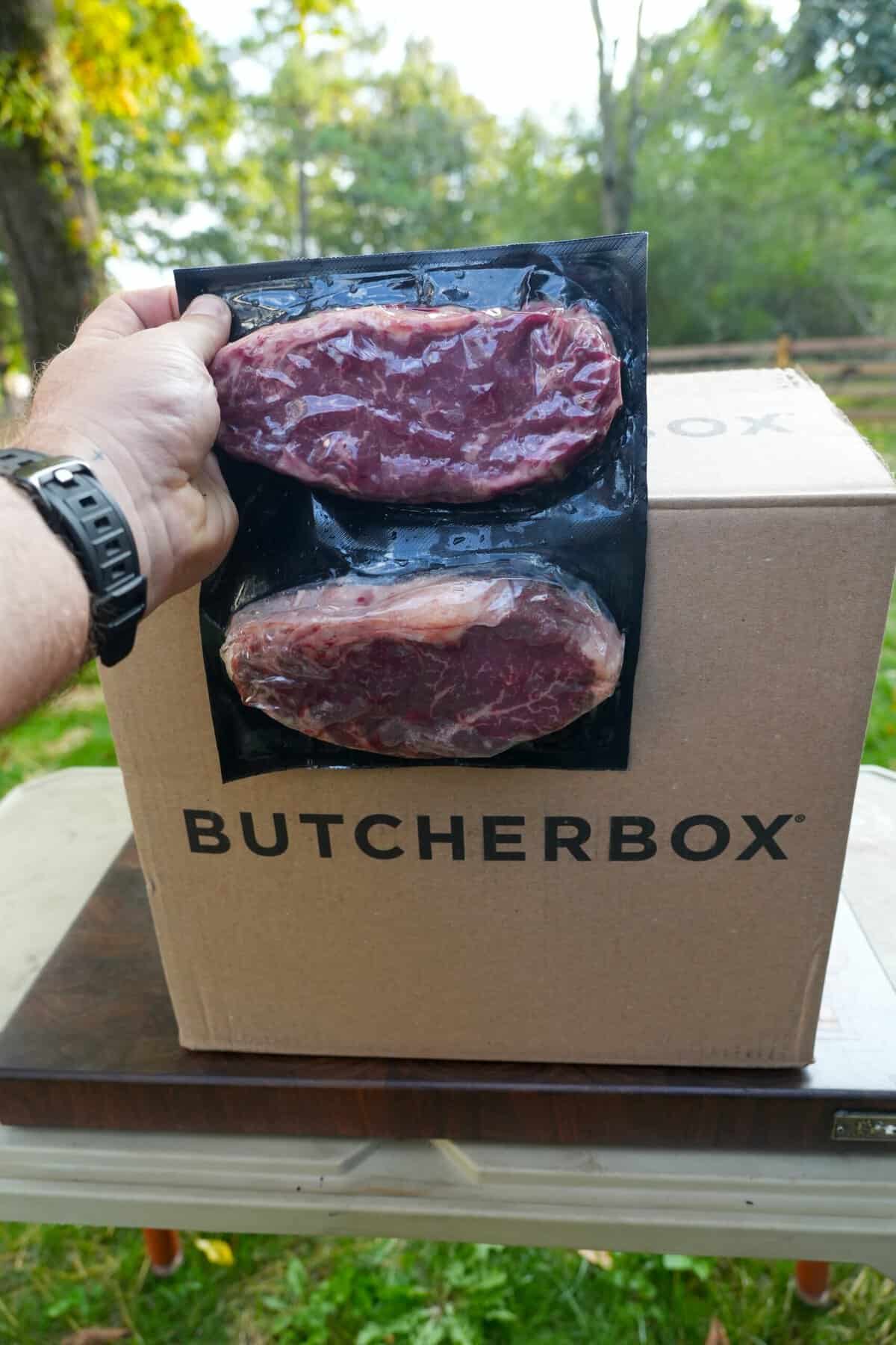 Derek Wolf holding up two raw steaks in plastic packaging, in front of a ButcherBox branded cardboard box
