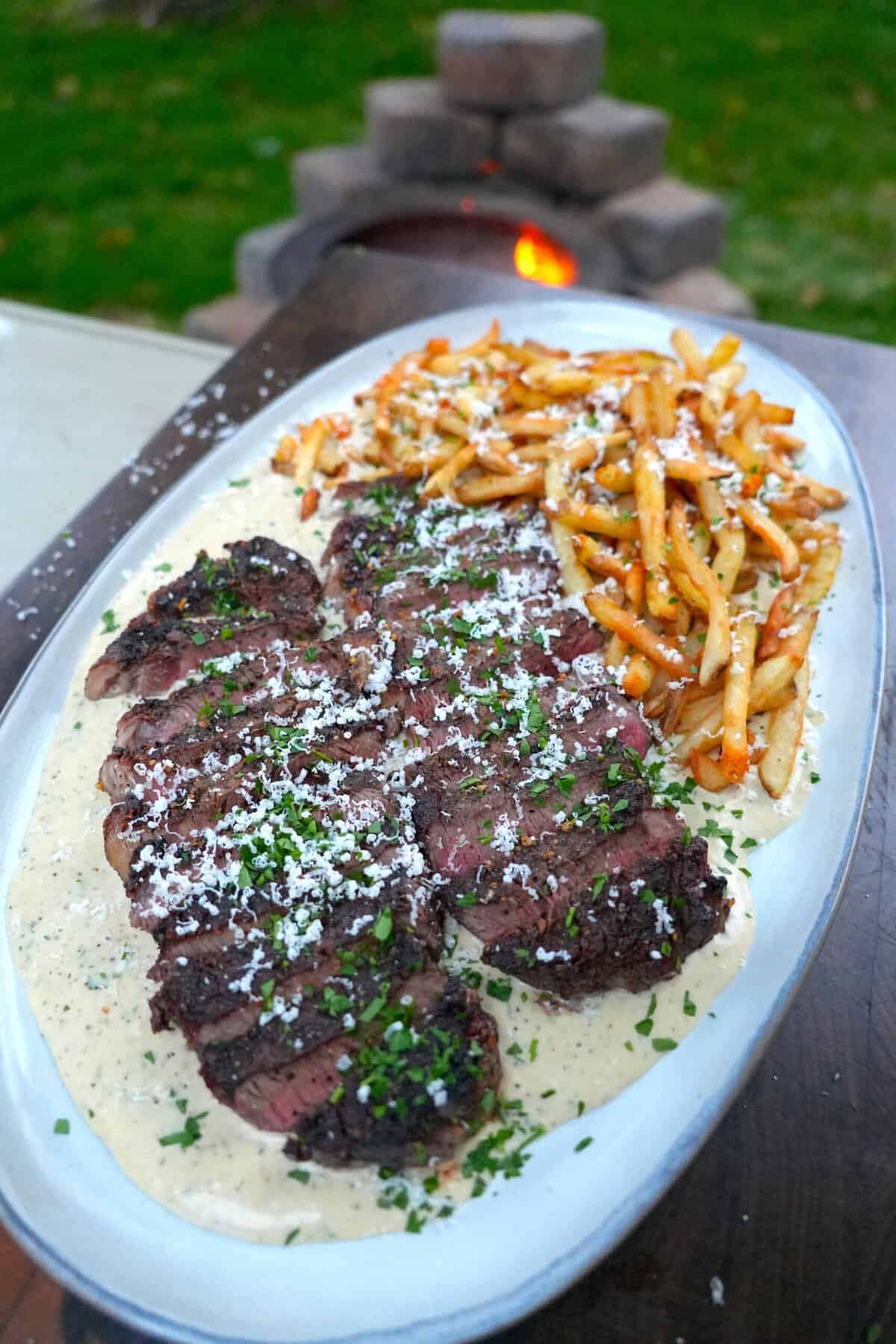 A finished plate of Alfredo steak and fries, with two steaks sitting in the sauce and garnished with Parmesan and parsley and the fries at the top of the plate