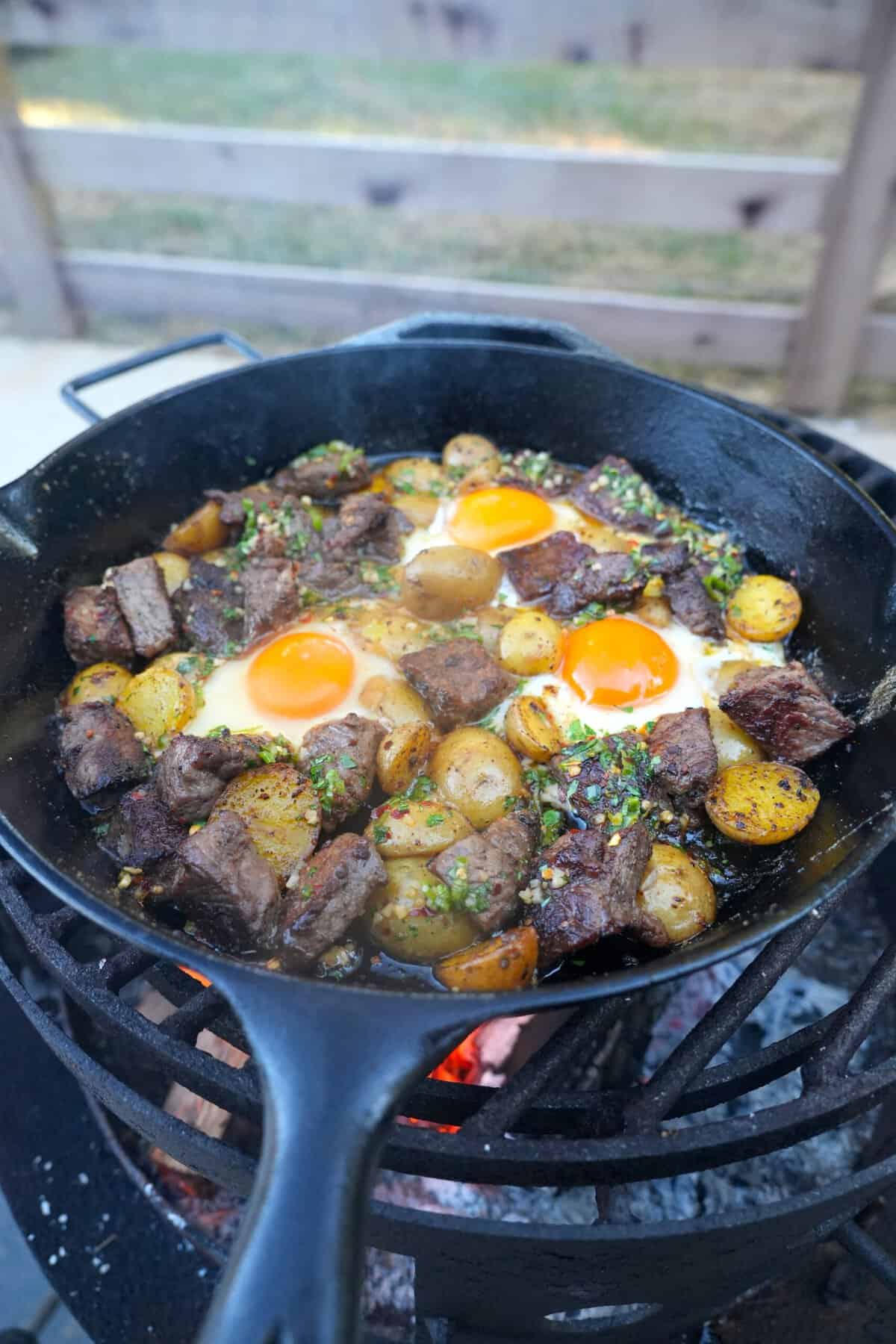 A wider shot of the steak and eggs cooking over the fire.