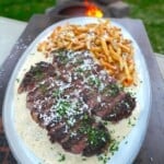 A finished plate of Alfredo steak and fries, on a white plate with garlic Parmesan sauce and a pile of homemade fries