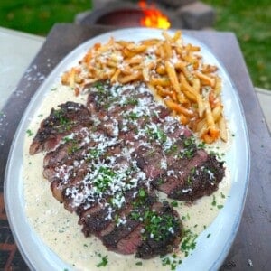 A finished plate of Alfredo steak and fries, on a white plate with garlic Parmesan sauce and a pile of homemade fries