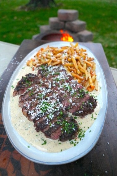 A finished plate of Alfredo steak and fries, on a white plate with garlic Parmesan sauce and a pile of homemade fries