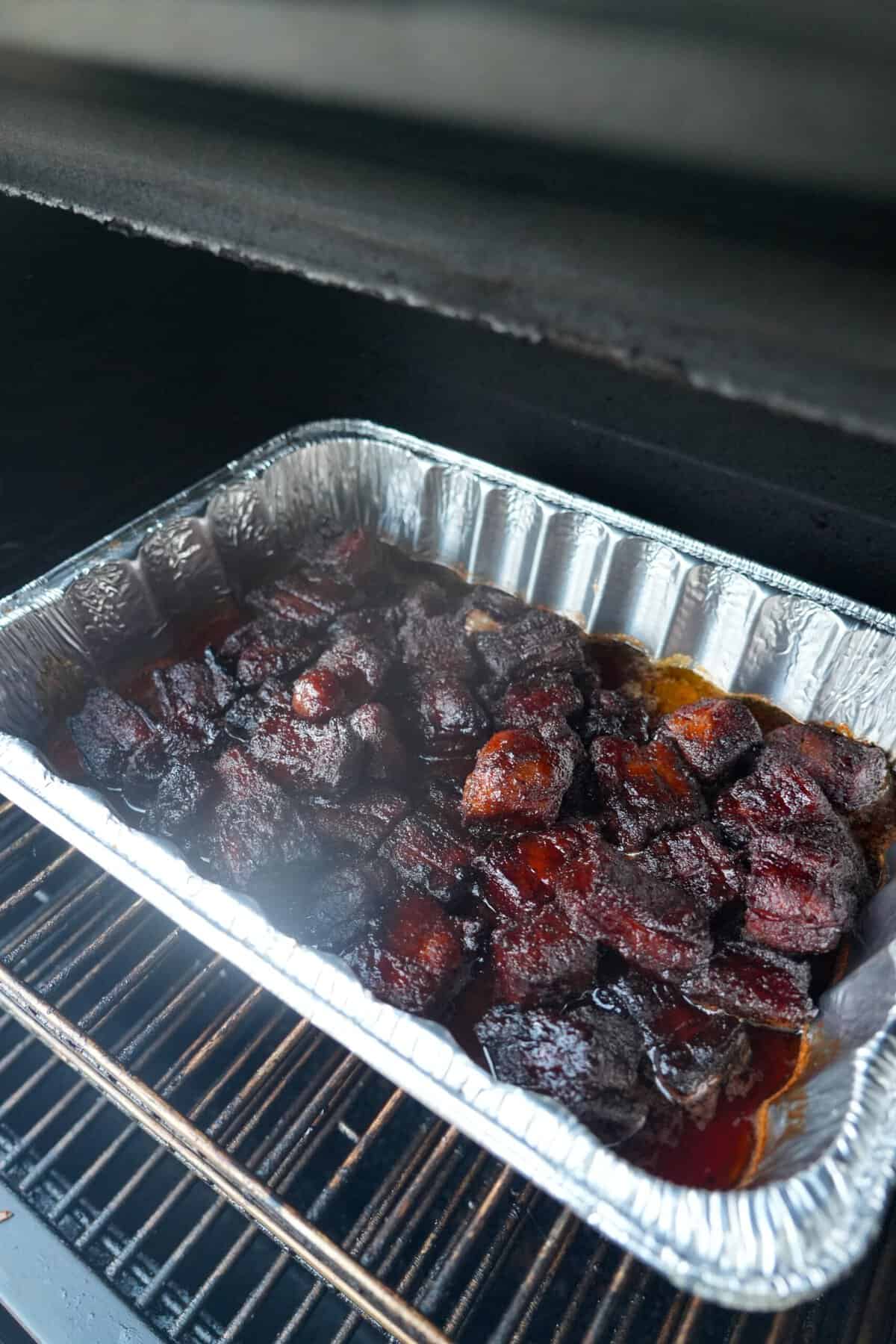 An aluminum pan on a smoker with cubes of pork belly burnt ends, basted with honey chipotle BBQ sauce