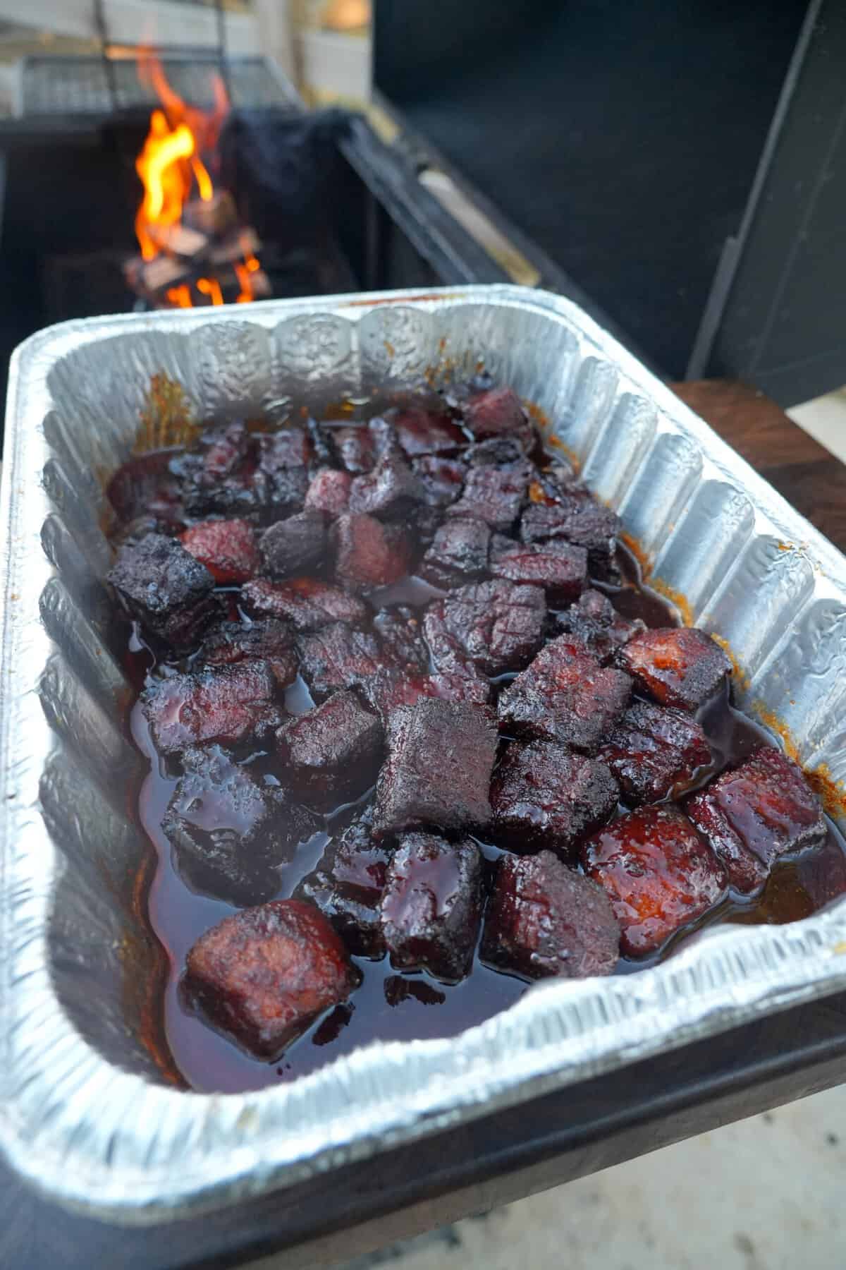 A close up of a foil pan on a smoker with cubes of pork belly burnt ends, cooking in a BBQ glaze