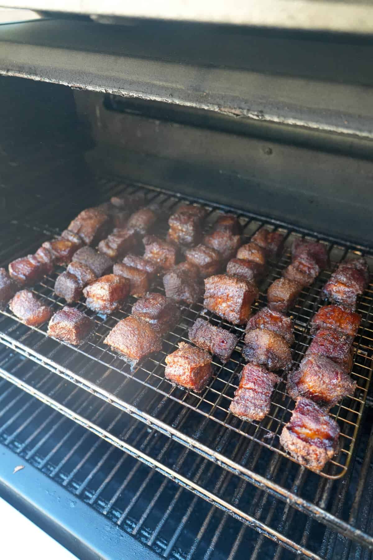 A lineup of pork belly cubes, cooking on smoker grates