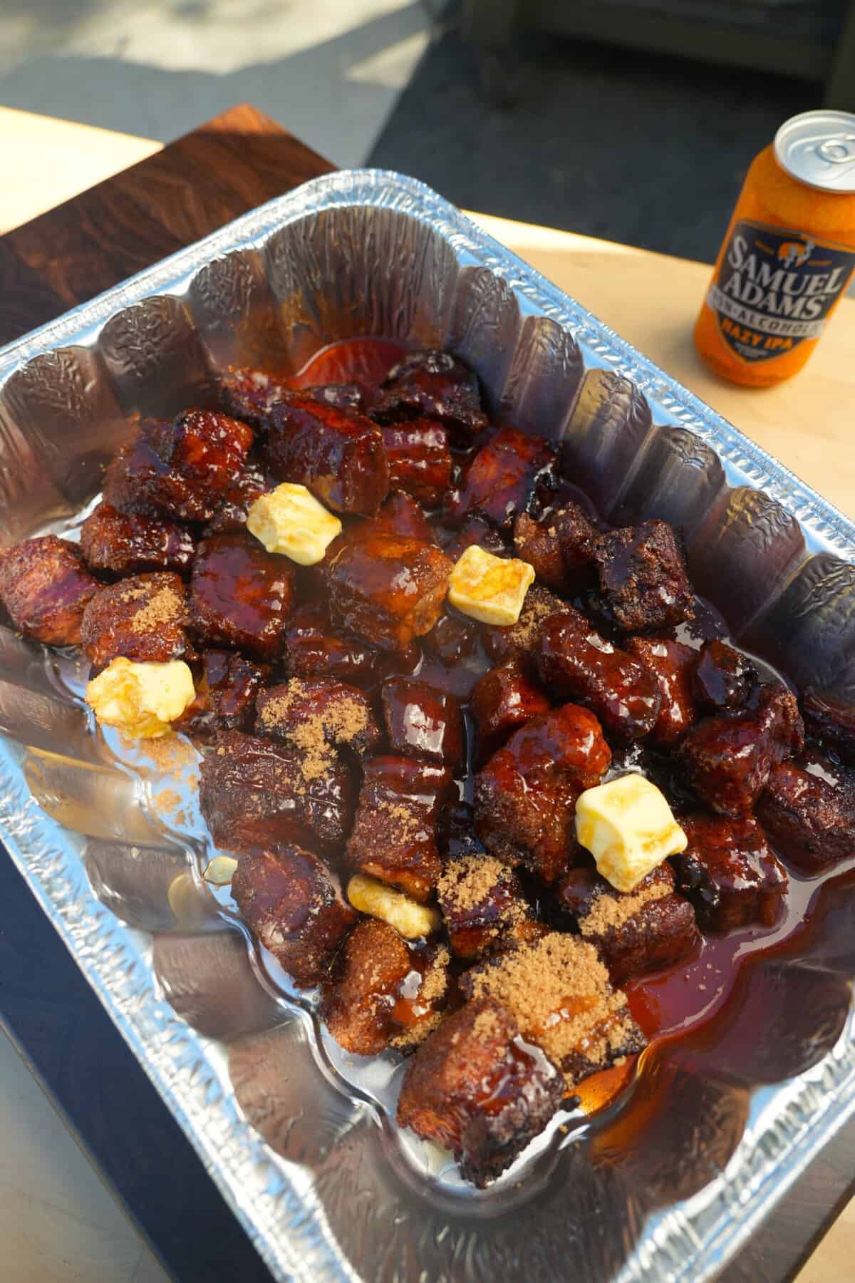 An aluminum pan filled with partially cooked pork belly burnt ends, with butter pads on top of the meat to start the crutch process