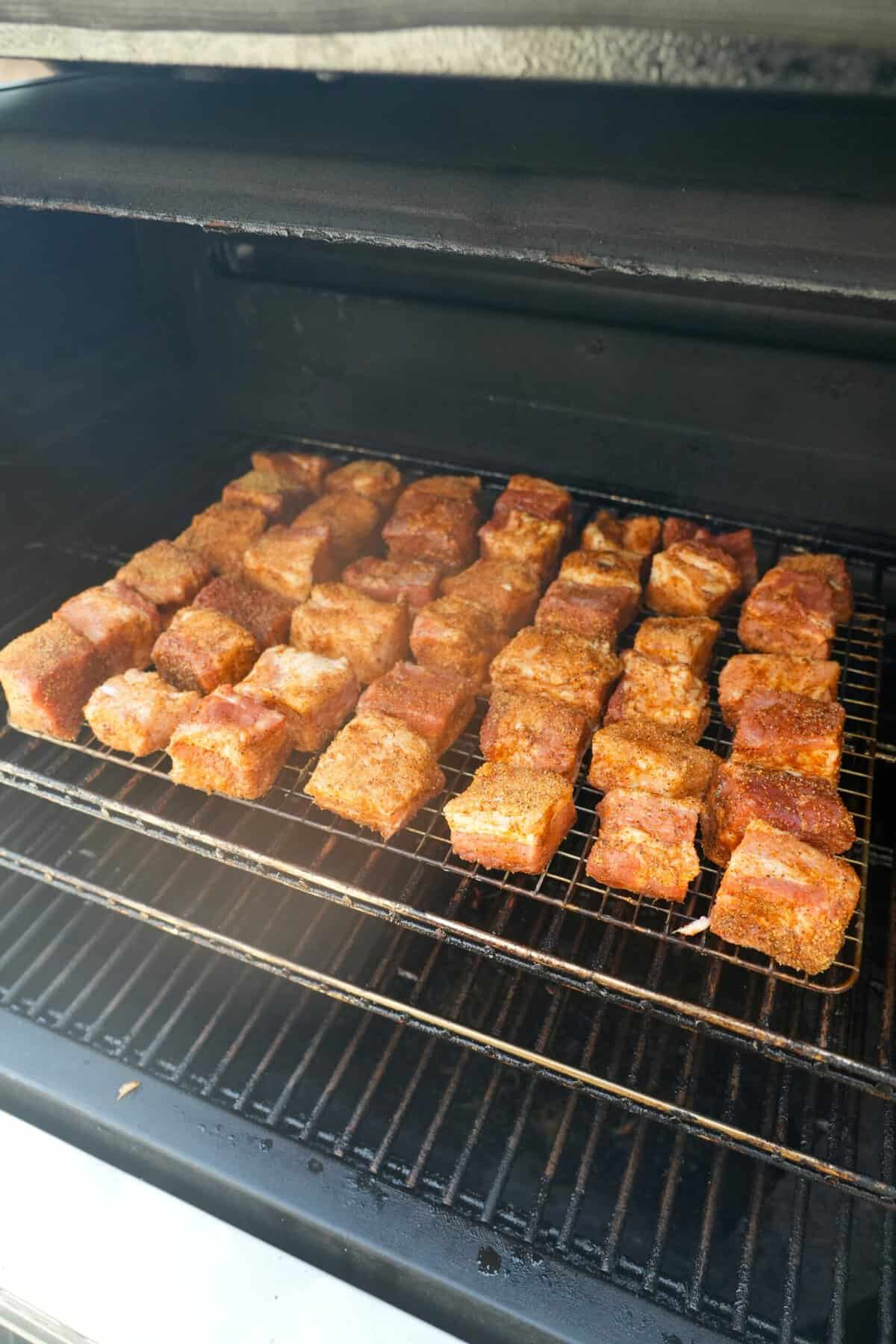 A lineup of raw, seasoned pork belly cubes on a smoker
