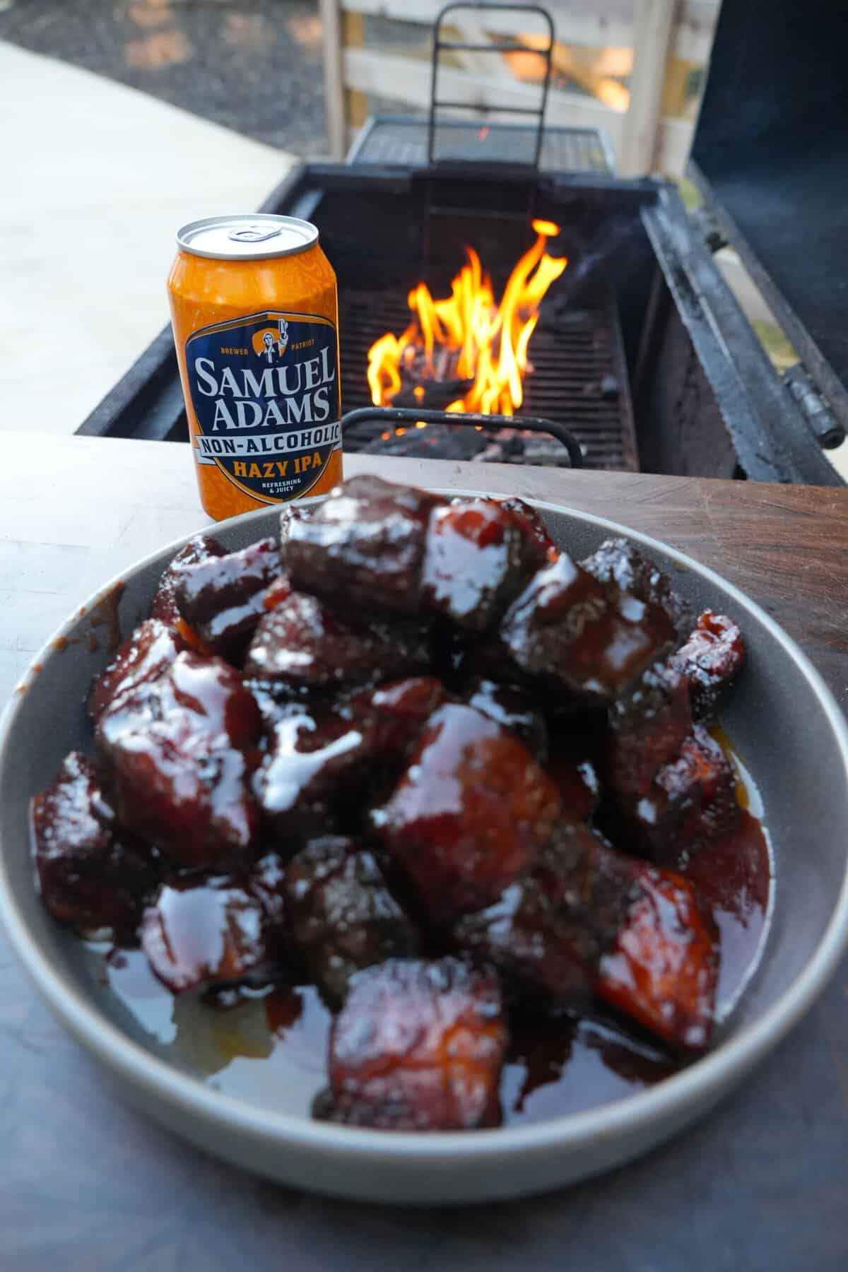 A plate of finished, glazed honey chipotle pork belly burnt ends, sitting on a table with a can of Samuel Adams Just the Haze and a grill fire in the background