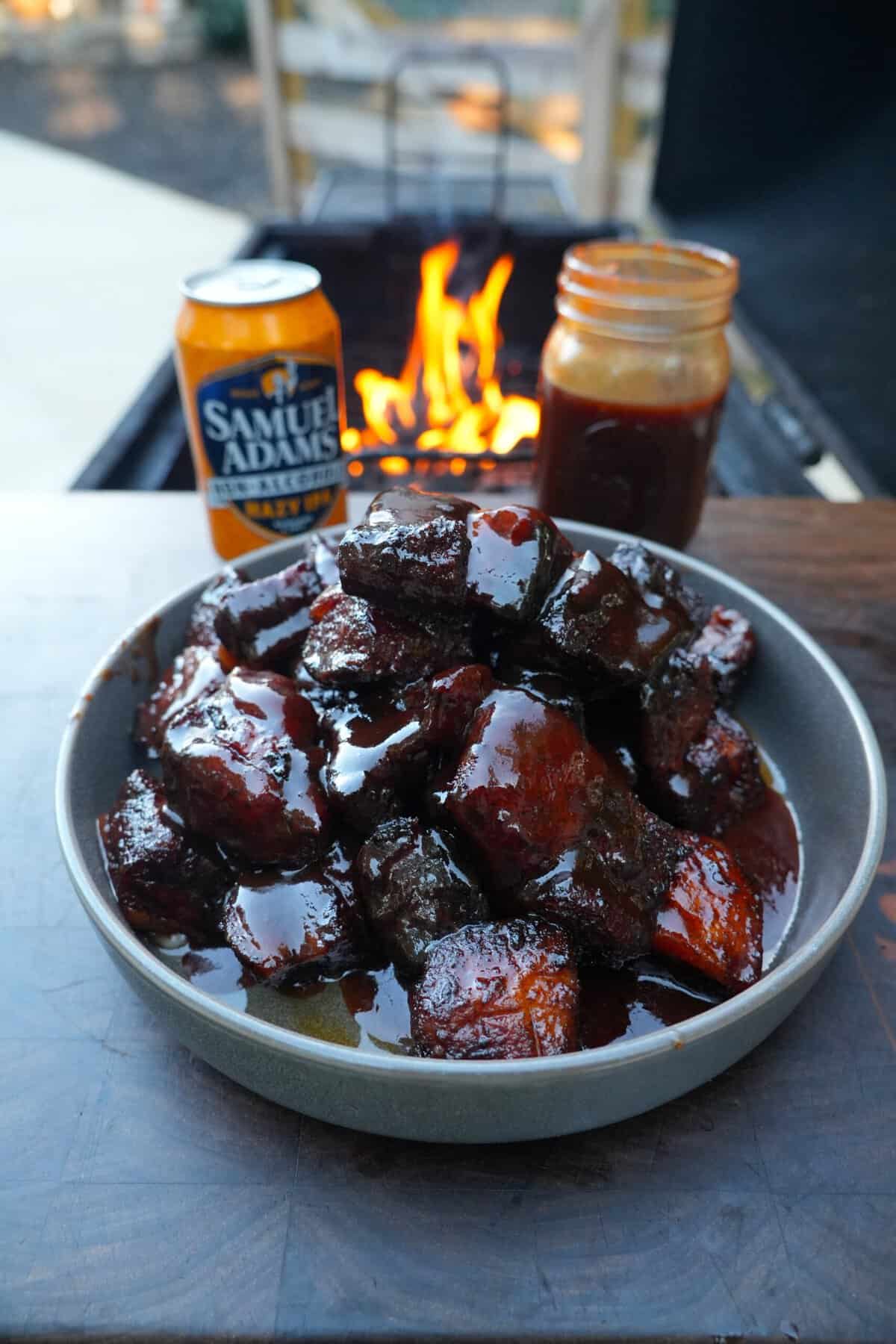 A plate of finished, glazed honey chipotle pork belly burnt ends, sitting on a table with a can of Samuel Adams Just the Haze, a jar of BBQ sauce and a grill fire in the background
