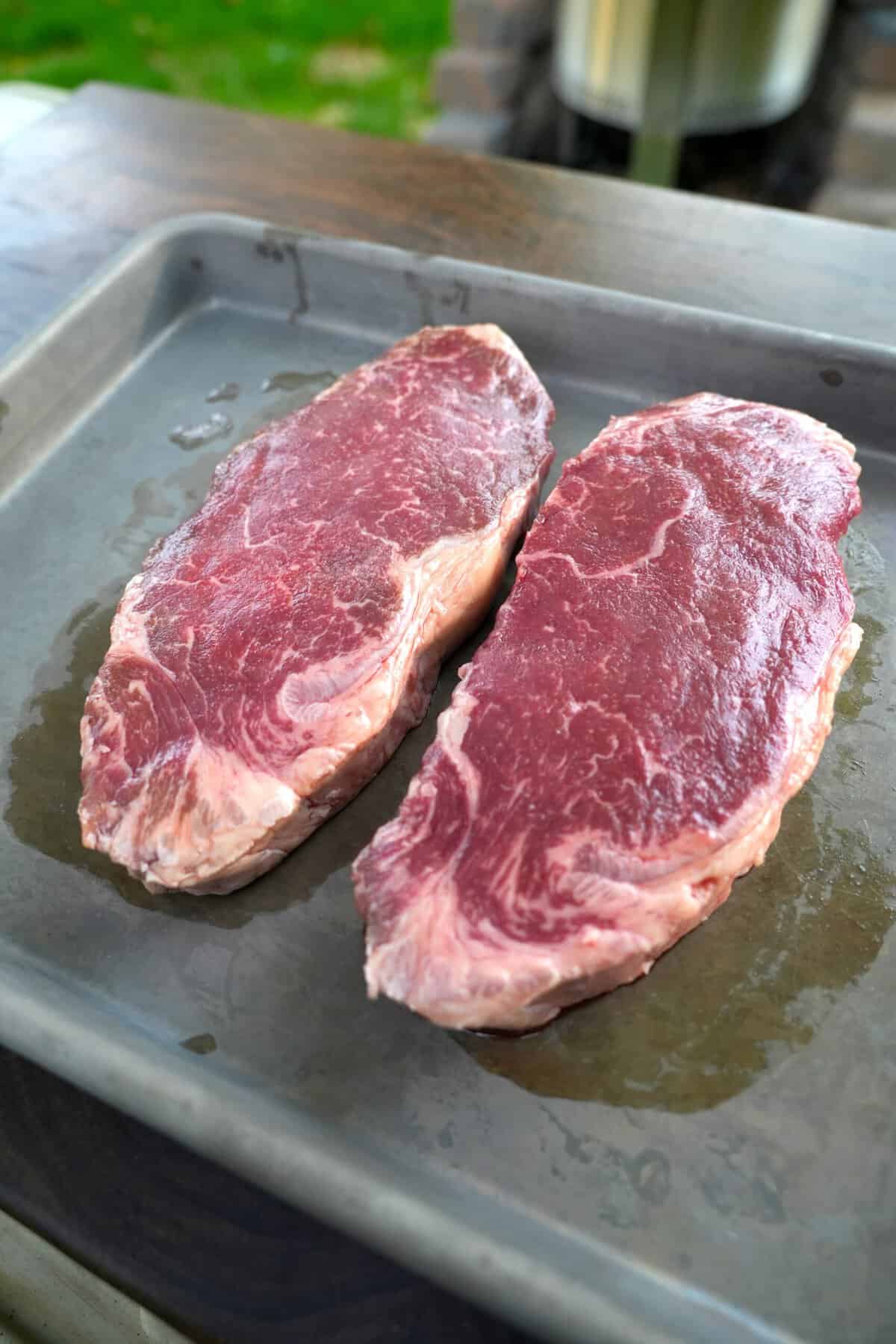 Two raw steaks sitting side by side on a baking sheet and coated with canola oil
